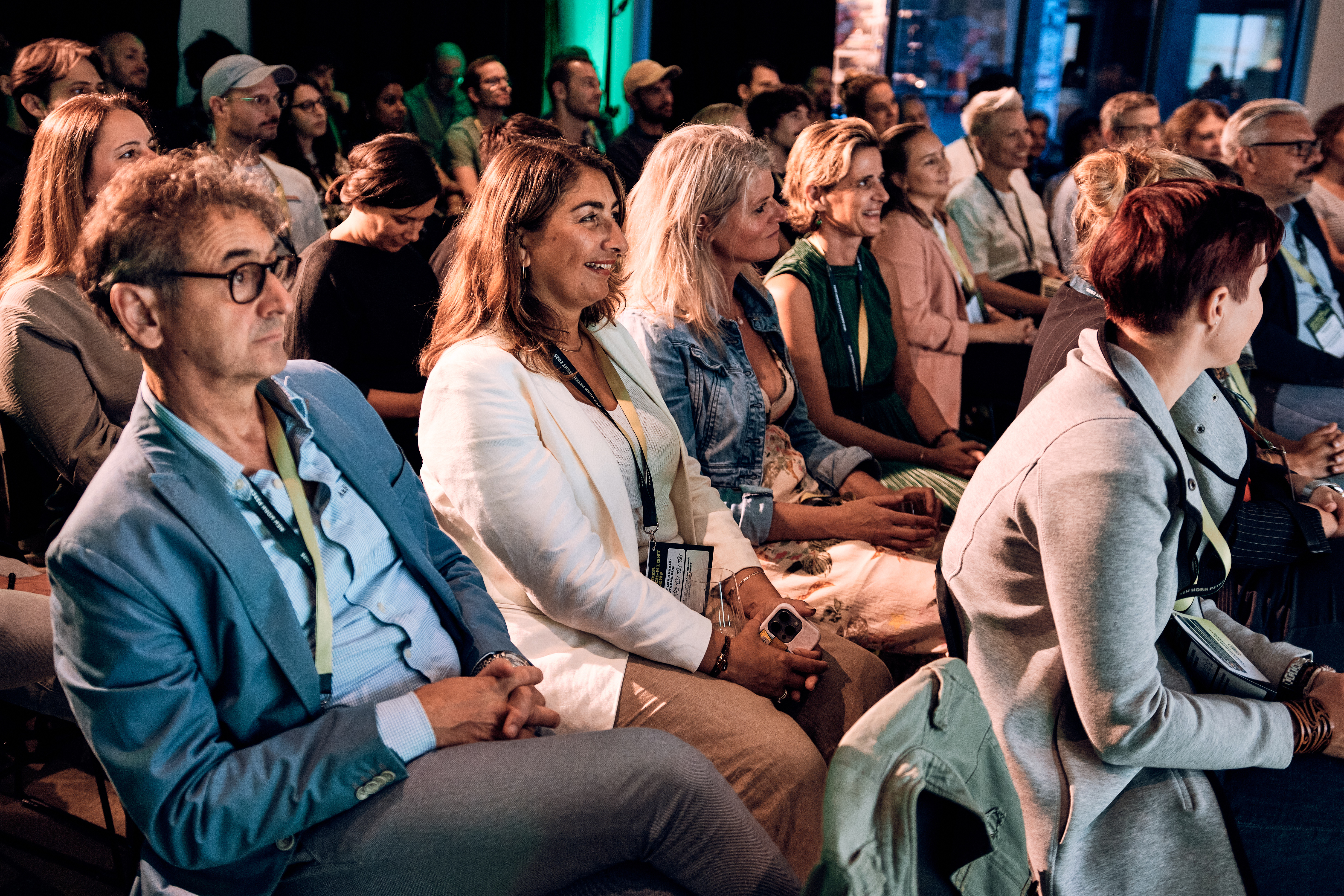 Erwachsene sitzen dicht gedrängt in Reihen und verfolgen aufmerksam eine Veranstaltung oder Präsentation in einem hellen, gut beleuchteten Raum.